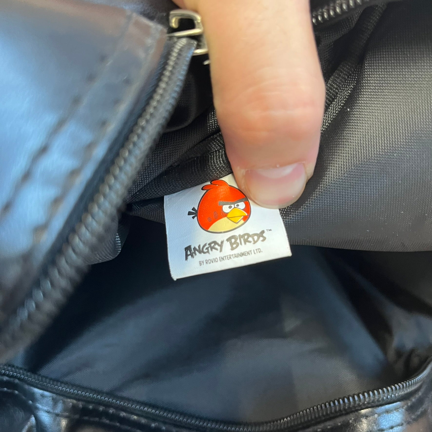 Angry Birds 2010s Black Bomb Bird Shoulder Bag is a black messenger-style bag made from a leather-like material. It features one main compartment with a smaller internal pocket. The front is designed with a large graphic of Bomb, the black Angry Birds character, shown with a screaming expression. The interior includes official Angry Birds 2009-2013 licensing.