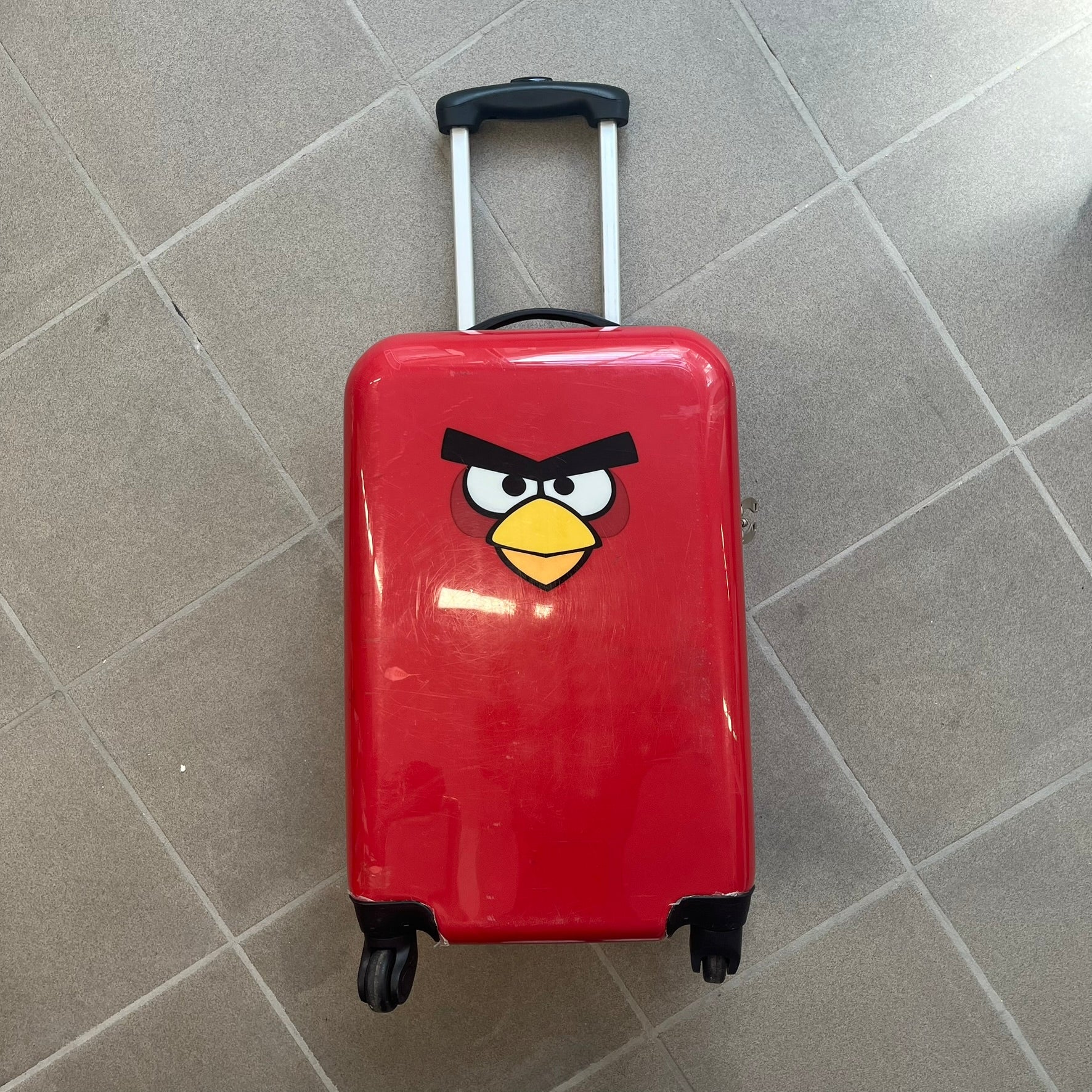 Angry Birds 2010s Red Bird Suitcase is a red rolling suitcase featuring the face of Red, the iconic Angry Birds character, on the front. It includes black wheels, a silver and black retractable handle, and an Angry Birds logo on the back. The interior features adjustable straps, zipped mesh compartments, and an all-over monogram lining with Angry Birds character patterns.