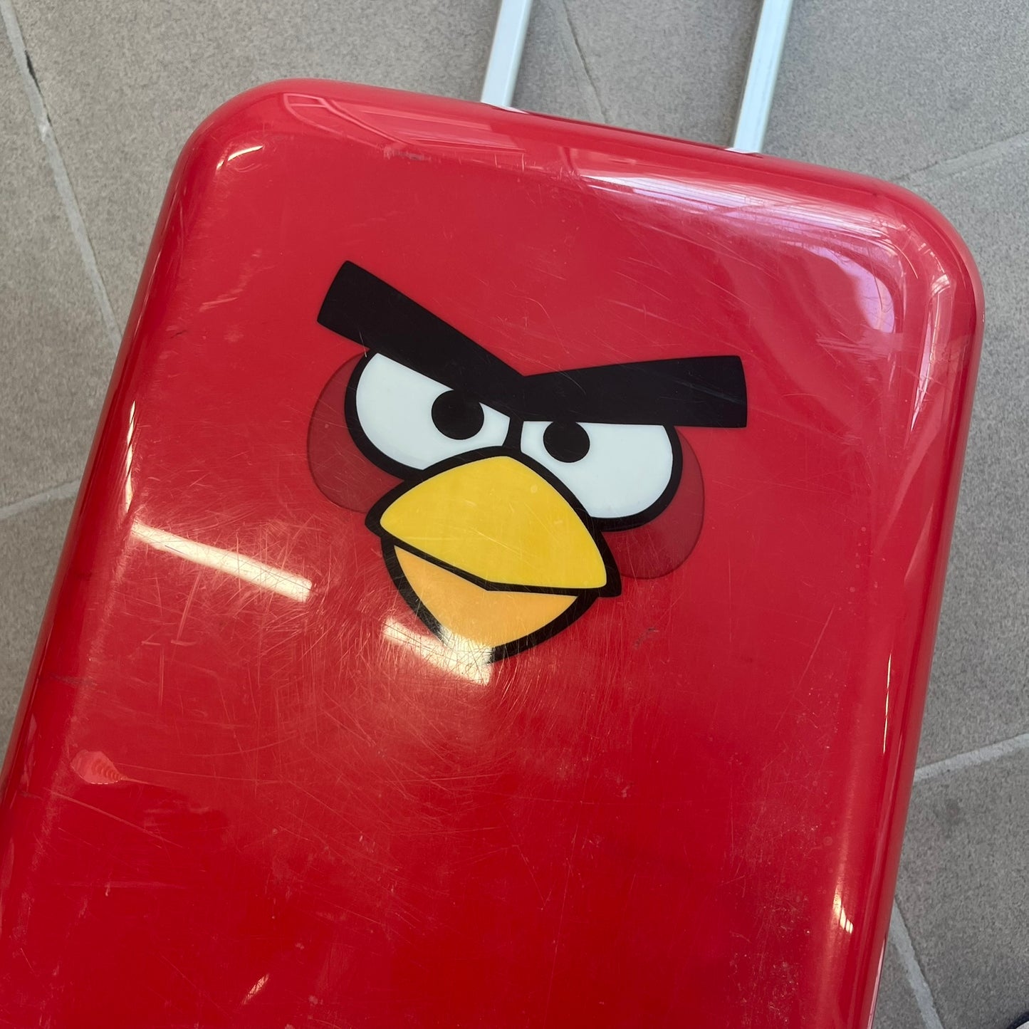 Angry Birds 2010s Red Bird Suitcase is a red rolling suitcase featuring the face of Red, the iconic Angry Birds character, on the front. It includes black wheels, a silver and black retractable handle, and an Angry Birds logo on the back. The interior features adjustable straps, zipped mesh compartments, and an all-over monogram lining with Angry Birds character patterns.