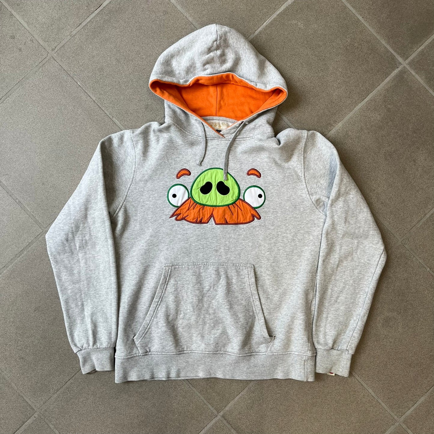 Angry Birds Three Star 2012 Foreman Pig Gray Hoodie features a grey hoodie design with a 3D-style front graphic of Foreman Pig, showing his recognizable eyes, nose, and mustache details. The back features a grey circular "Three Star" Angry Birds graphic, while the inside of the hood is finished with an orange lining.