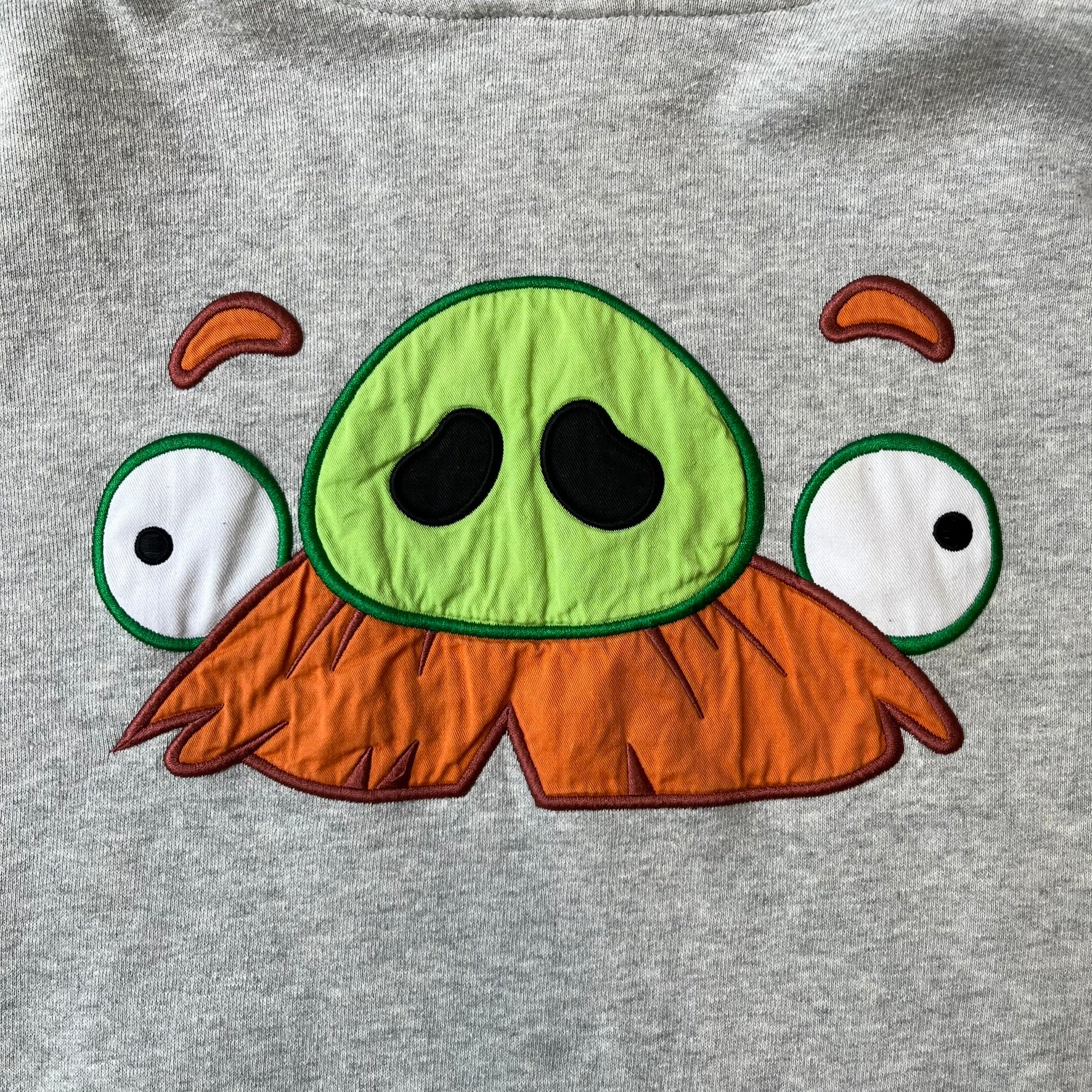 Angry Birds Three Star 2012 Foreman Pig Gray Hoodie features a grey hoodie design with a 3D-style front graphic of Foreman Pig, showing his recognizable eyes, nose, and mustache details. The back features a grey circular "Three Star" Angry Birds graphic, while the inside of the hood is finished with an orange lining.