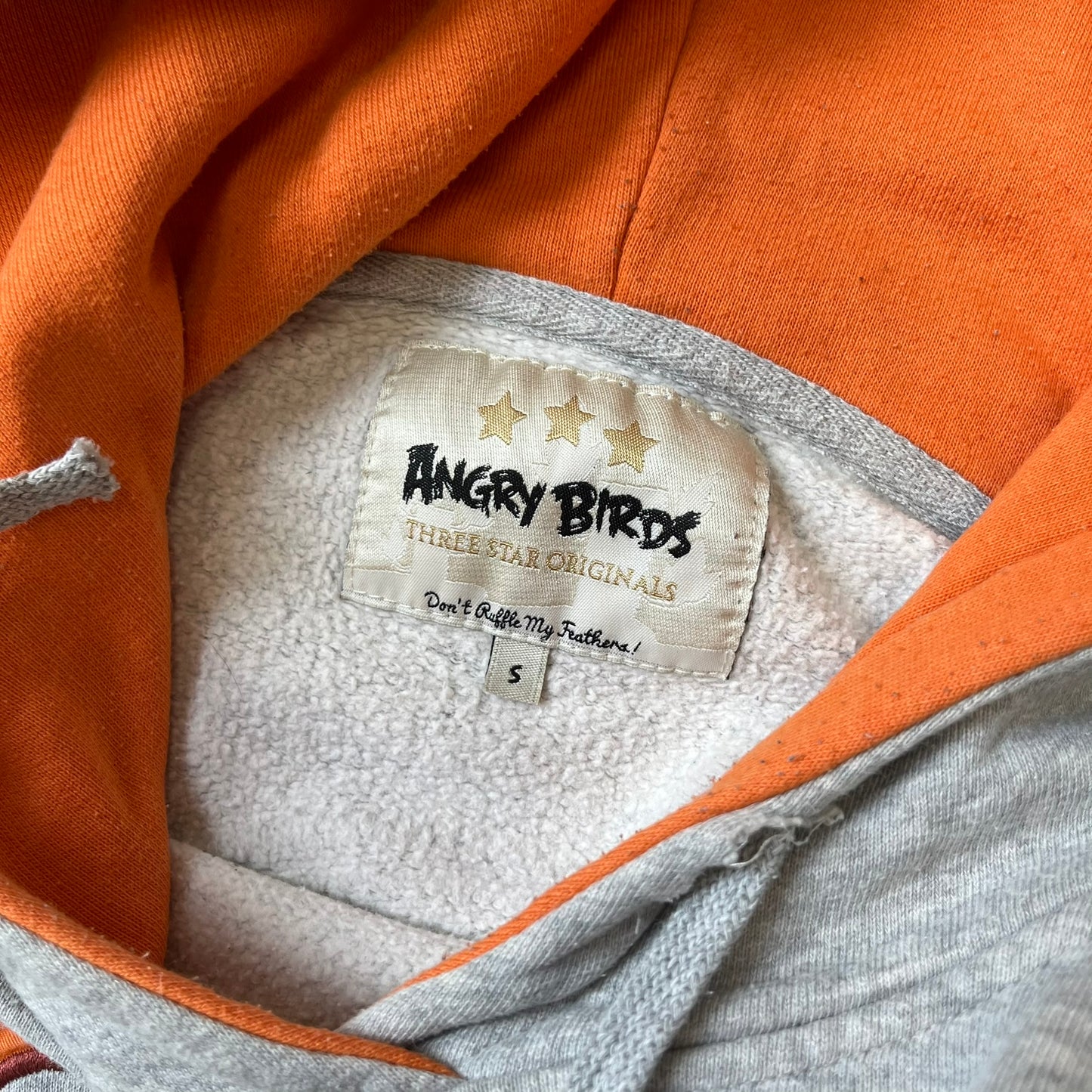 Angry Birds Three Star 2012 Foreman Pig Gray Hoodie features a grey hoodie design with a 3D-style front graphic of Foreman Pig, showing his recognizable eyes, nose, and mustache details. The back features a grey circular "Three Star" Angry Birds graphic, while the inside of the hood is finished with an orange lining.