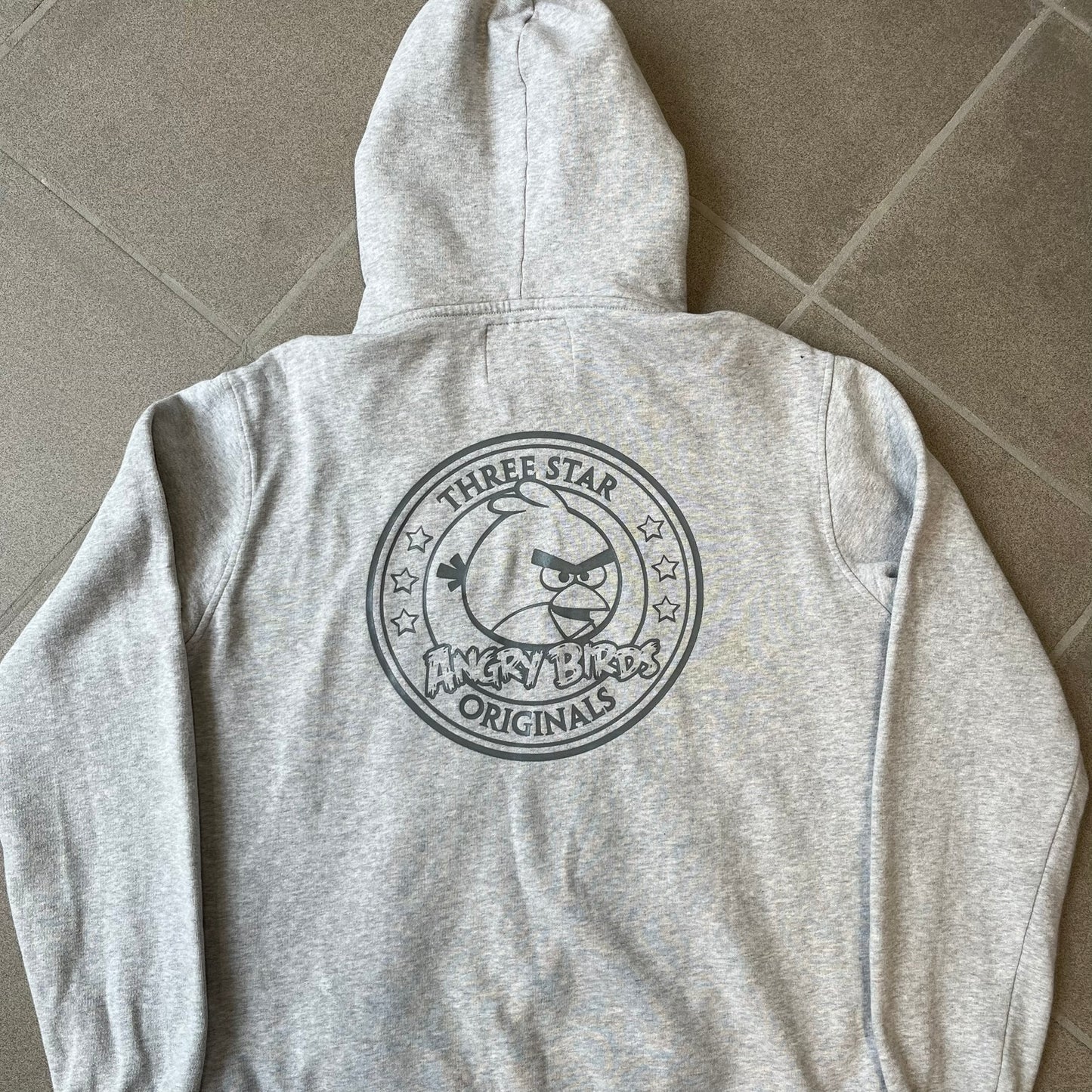 Angry Birds Three Star 2012 Foreman Pig Gray Hoodie features a grey hoodie design with a 3D-style front graphic of Foreman Pig, showing his recognizable eyes, nose, and mustache details. The back features a grey circular "Three Star" Angry Birds graphic, while the inside of the hood is finished with an orange lining.