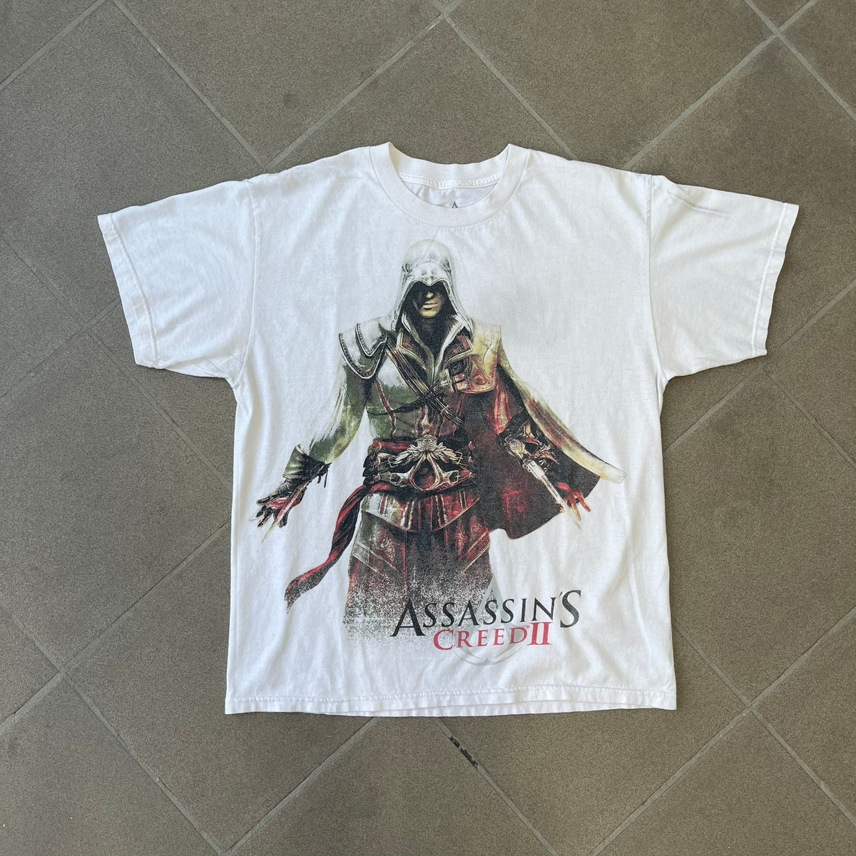 Assassin's Creed 2 Promo 2010 White Tee features a white 100% cotton construction with a large colorful graphic of Ezio Auditore da Firenze on the front. The Assassin's Creed 2 logo appears underneath the character, finished with a licensed neck tag.
