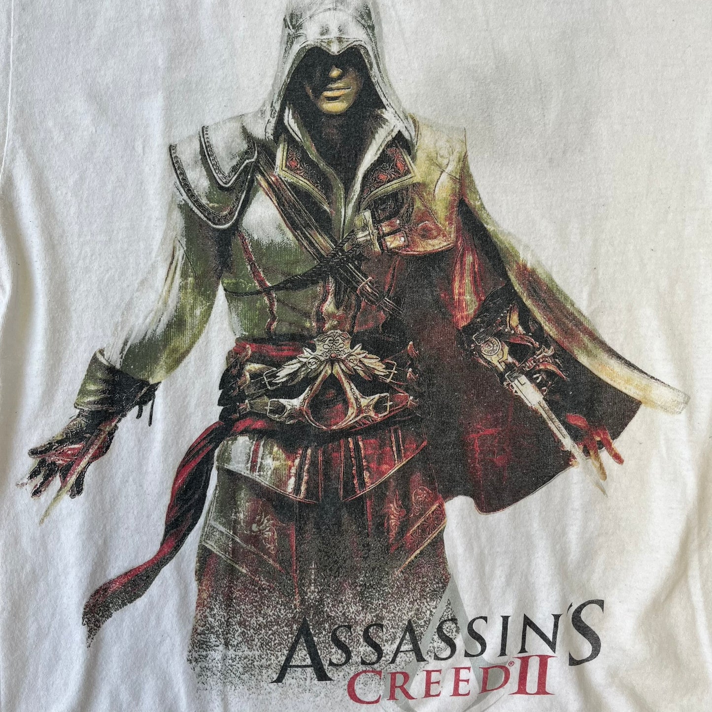 Assassin's Creed 2 Promo 2010 White Tee features a white 100% cotton construction with a large colorful graphic of Ezio Auditore da Firenze on the front. The Assassin's Creed 2 logo appears underneath the character, finished with a licensed neck tag.
