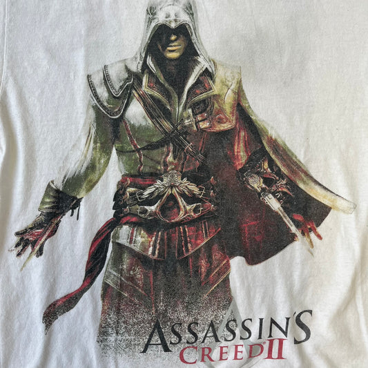 Assassin's Creed 2 Promo 2010 White Tee features a white 100% cotton construction with a large colorful graphic of Ezio Auditore da Firenze on the front. The Assassin's Creed 2 logo appears underneath the character, finished with a licensed neck tag.
