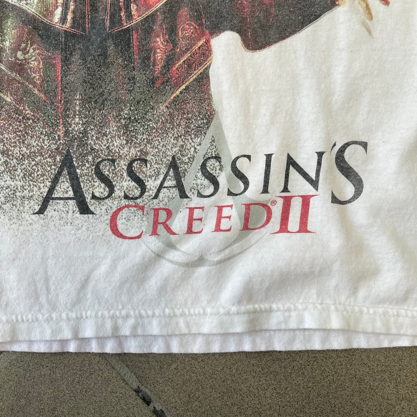 Assassin's Creed 2 Promo 2010 White Tee features a white 100% cotton construction with a large colorful graphic of Ezio Auditore da Firenze on the front. The Assassin's Creed 2 logo appears underneath the character, finished with a licensed neck tag.
