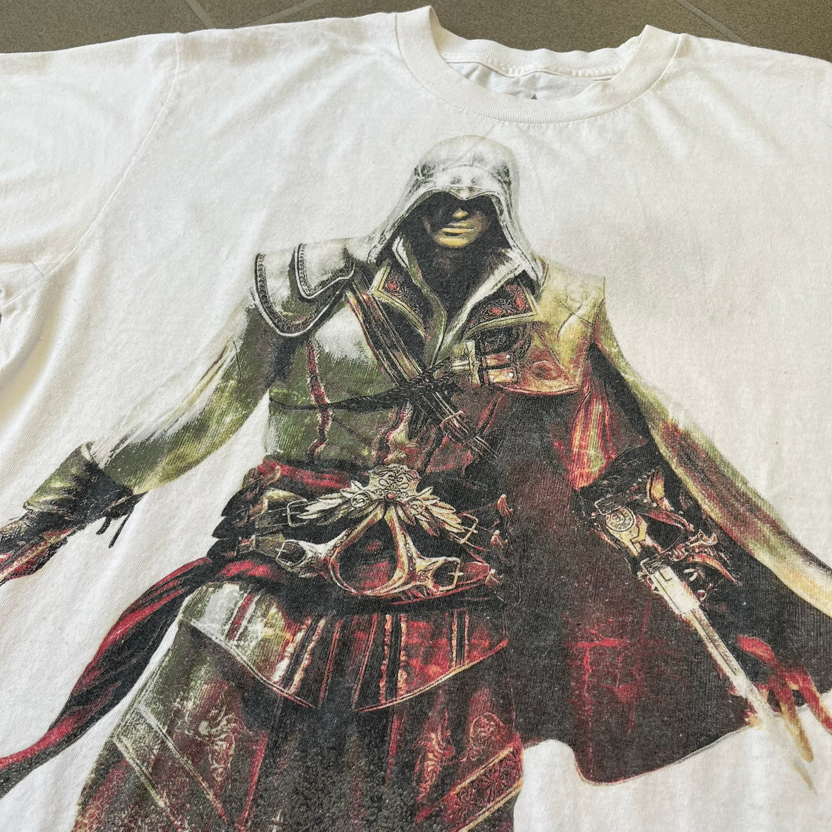Assassin's Creed 2 Promo 2010 White Tee features a white 100% cotton construction with a large colorful graphic of Ezio Auditore da Firenze on the front. The Assassin's Creed 2 logo appears underneath the character, finished with a licensed neck tag.
