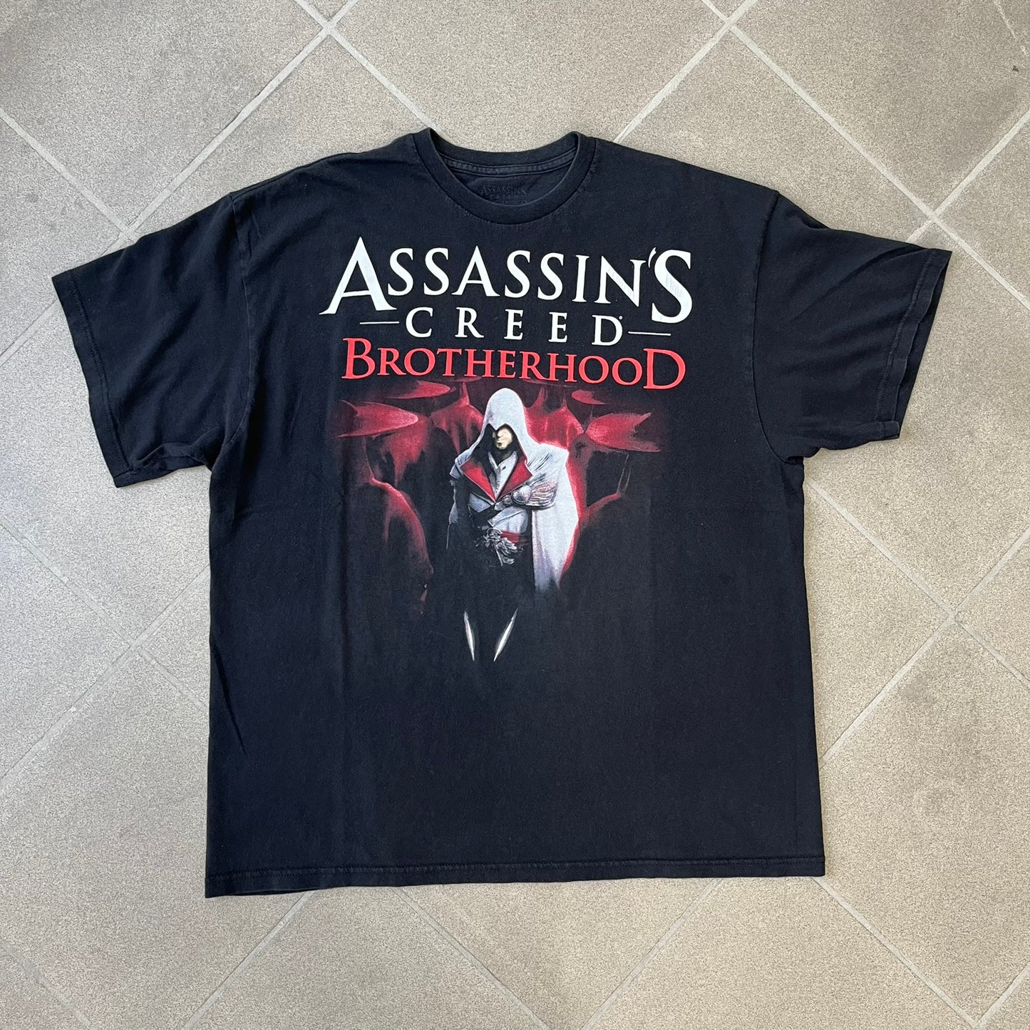 Assassin's Creed Brotherhood 2010 Black Promo Tee features a black construction with the Assassin's Creed Brotherhood cover printed on the front, showing Ezio Auditore with the large white and red text above him. Finished with a washed-off licensed neck tag