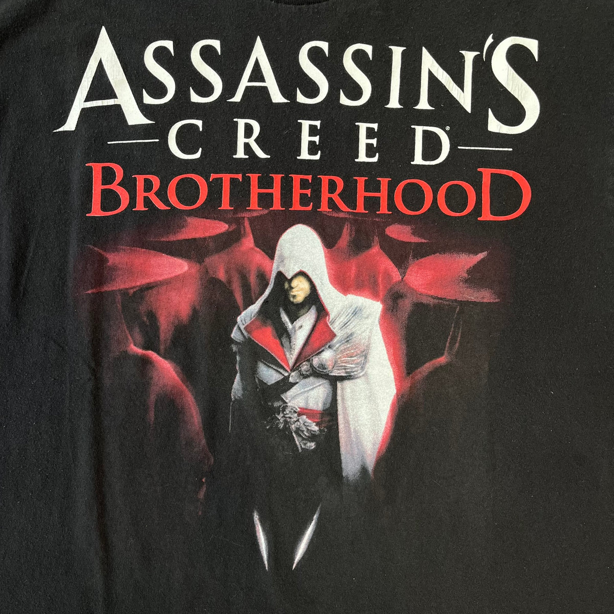Assassin's Creed Brotherhood 2010 Black Promo Tee features a black construction with the Assassin's Creed Brotherhood cover printed on the front, showing Ezio Auditore with the large white and red text above him. Finished with a washed-off licensed neck tag