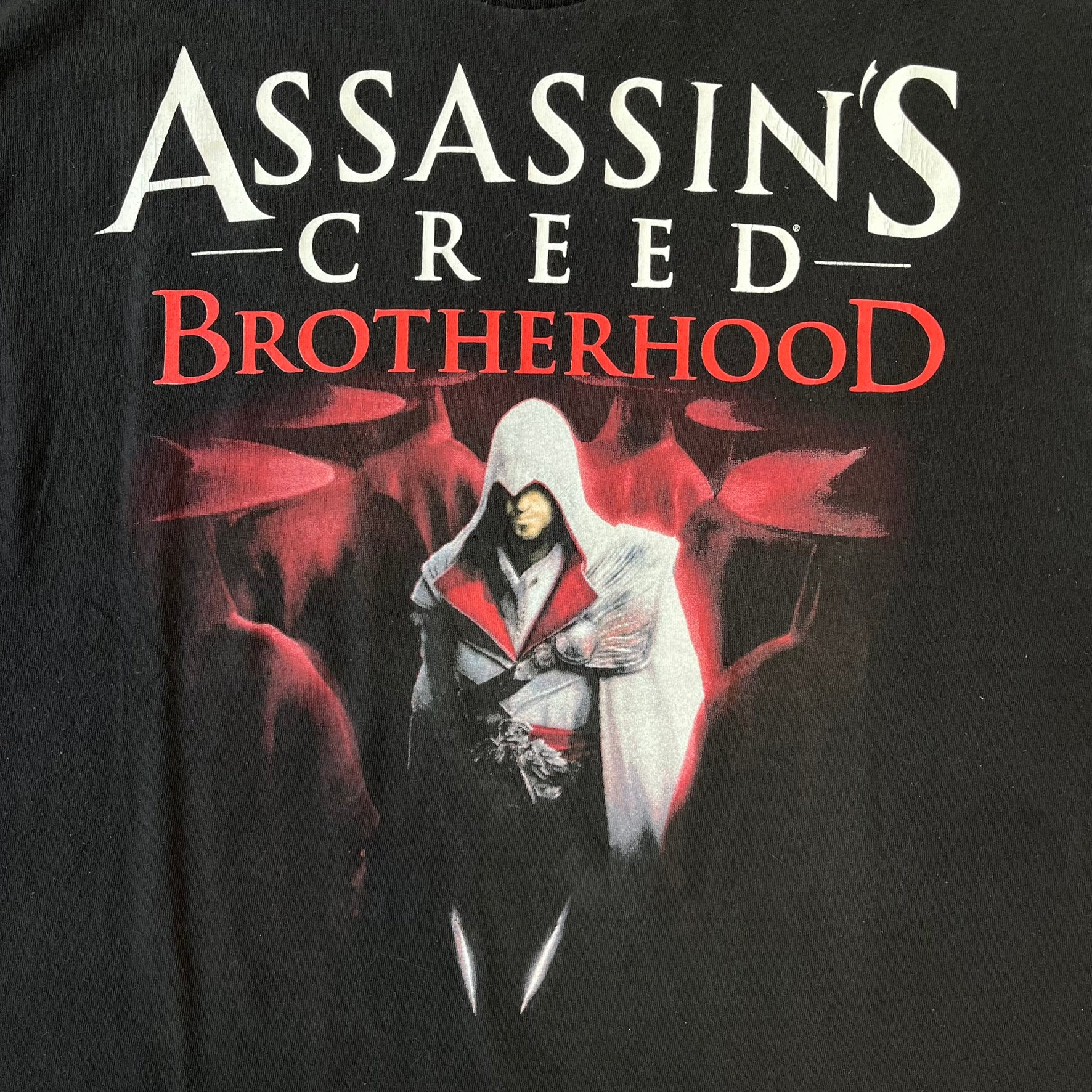 Assassin's Creed Brotherhood 2010 Black Promo Tee features a black construction with the Assassin's Creed Brotherhood cover printed on the front, showing Ezio Auditore with the large white and red text above him. Finished with a washed-off licensed neck tag