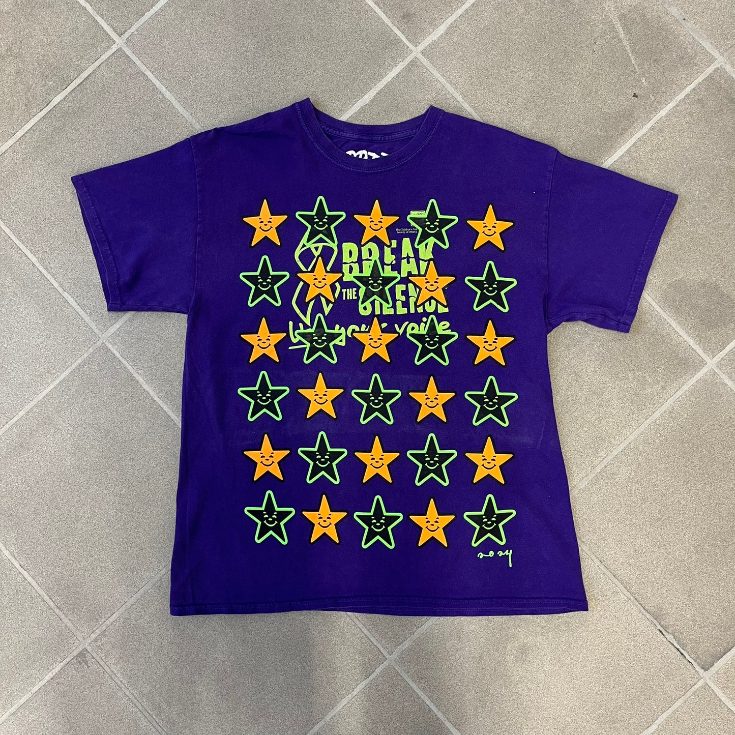 Asspizza 2024 Halloween Stars Purple Tee is a purple T-shirt featuring an all-over star pattern with orange and black stars outlined in green. Each star has a smiling face graphic in the center. The shirt includes a Babbitt 2024 neck tag and is printed on a Children’s Aid blank.
