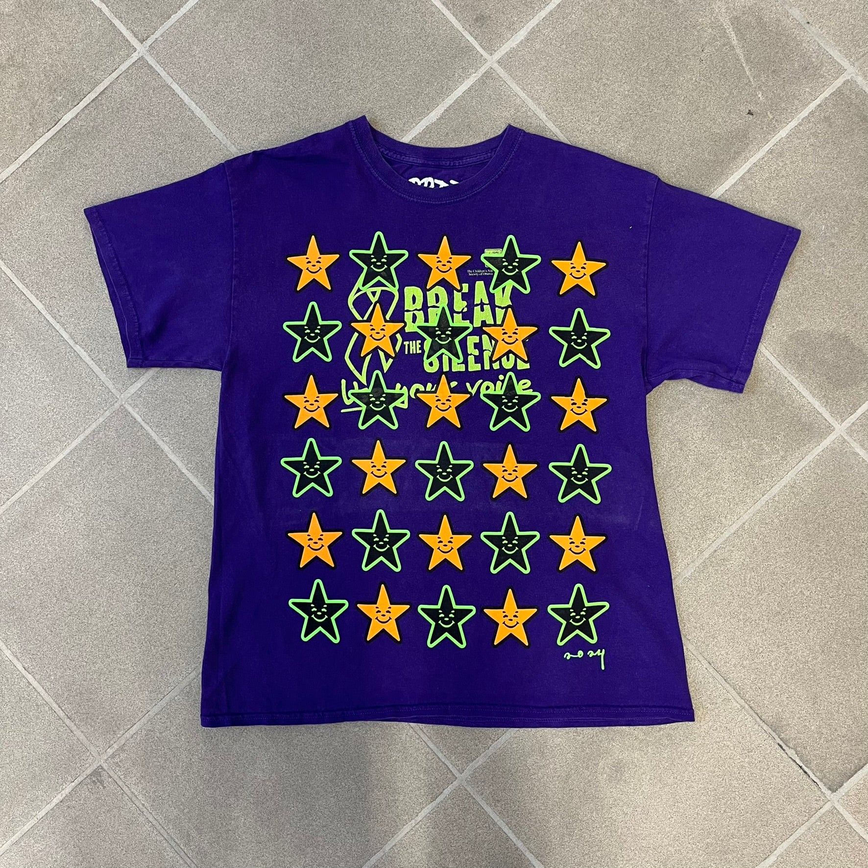 Asspizza 2024 Halloween Stars Purple Tee is a purple T-shirt featuring an all-over star pattern with orange and black stars outlined in green. Each star has a smiling face graphic in the center. The shirt includes a Babbitt 2024 neck tag and is printed on a Children’s Aid blank.
