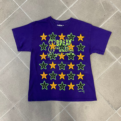 Asspizza 2024 Halloween Stars Purple Tee is a purple T-shirt featuring an all-over star pattern with orange and black stars outlined in green. Each star has a smiling face graphic in the center. The shirt includes a Babbitt 2024 neck tag and is printed on a Children’s Aid blank.
