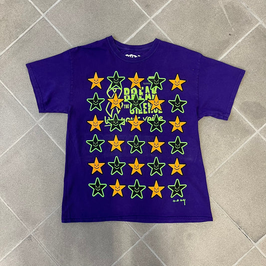 Asspizza 2024 Halloween Stars Purple Tee is a purple T-shirt featuring an all-over star pattern with orange and black stars outlined in green. Each star has a smiling face graphic in the center. The shirt includes a Babbitt 2024 neck tag and is printed on a Children’s Aid blank.
