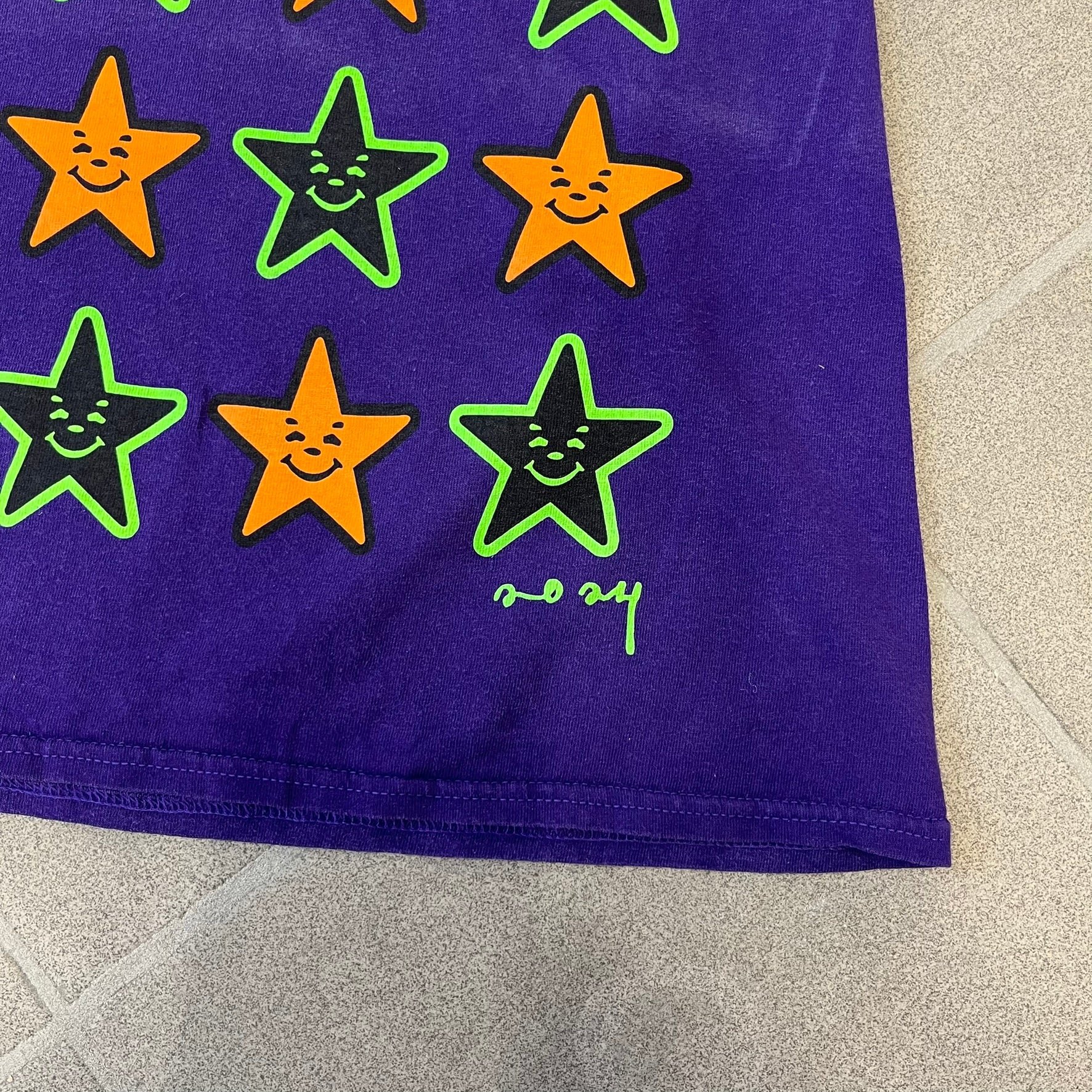 Asspizza 2024 Halloween Stars Purple Tee is a purple T-shirt featuring an all-over star pattern with orange and black stars outlined in green. Each star has a smiling face graphic in the center. The shirt includes a Babbitt 2024 neck tag and is printed on a Children’s Aid blank.
