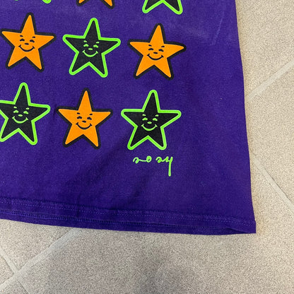 Asspizza 2024 Halloween Stars Purple Tee is a purple T-shirt featuring an all-over star pattern with orange and black stars outlined in green. Each star has a smiling face graphic in the center. The shirt includes a Babbitt 2024 neck tag and is printed on a Children’s Aid blank.
