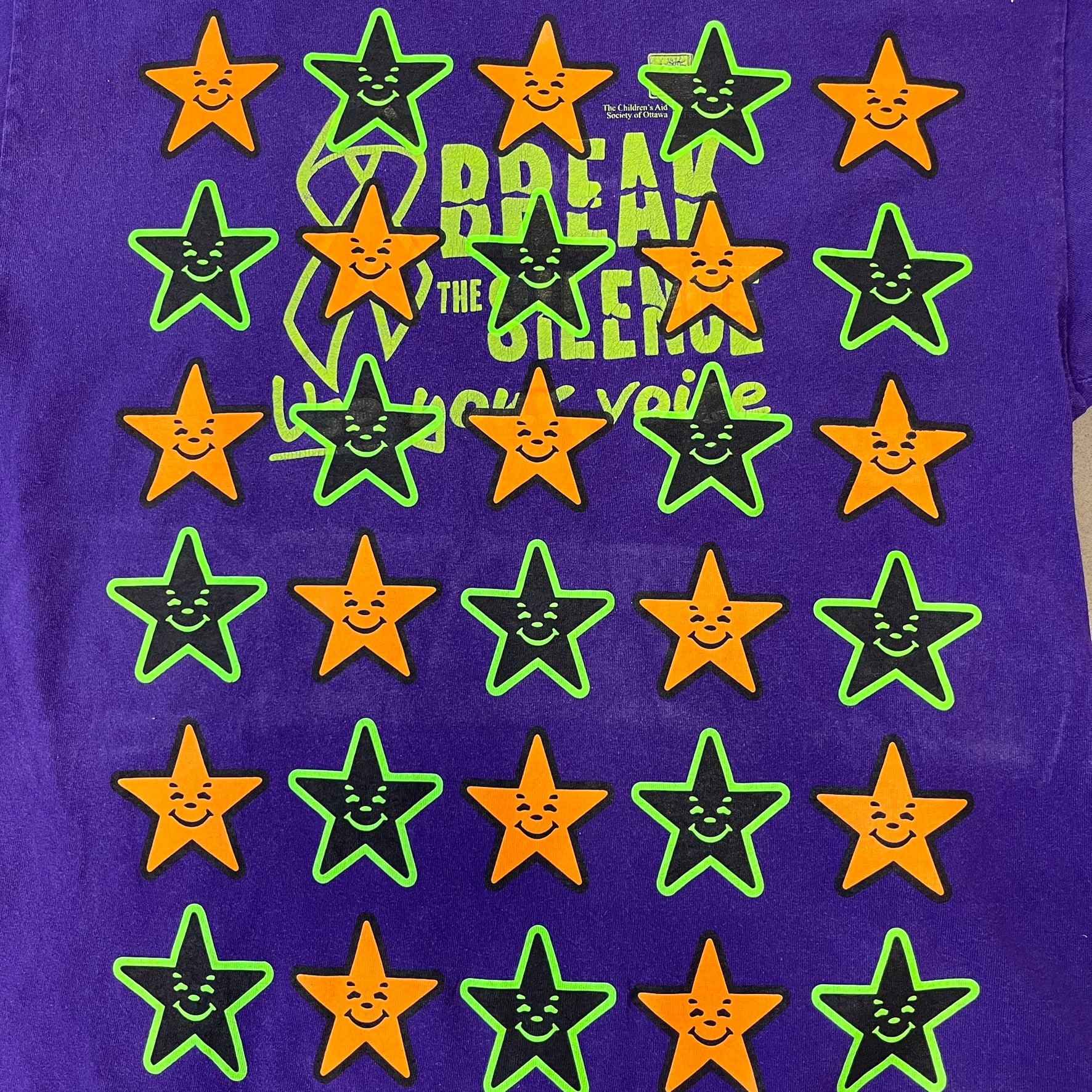 Asspizza 2024 Halloween Stars Purple Tee is a purple T-shirt featuring an all-over star pattern with orange and black stars outlined in green. Each star has a smiling face graphic in the center. The shirt includes a Babbitt 2024 neck tag and is printed on a Children’s Aid blank.
