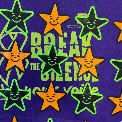 Asspizza 2024 Halloween Stars Purple Tee is a purple T-shirt featuring an all-over star pattern with orange and black stars outlined in green. Each star has a smiling face graphic in the center. The shirt includes a Babbitt 2024 neck tag and is printed on a Children’s Aid blank.
