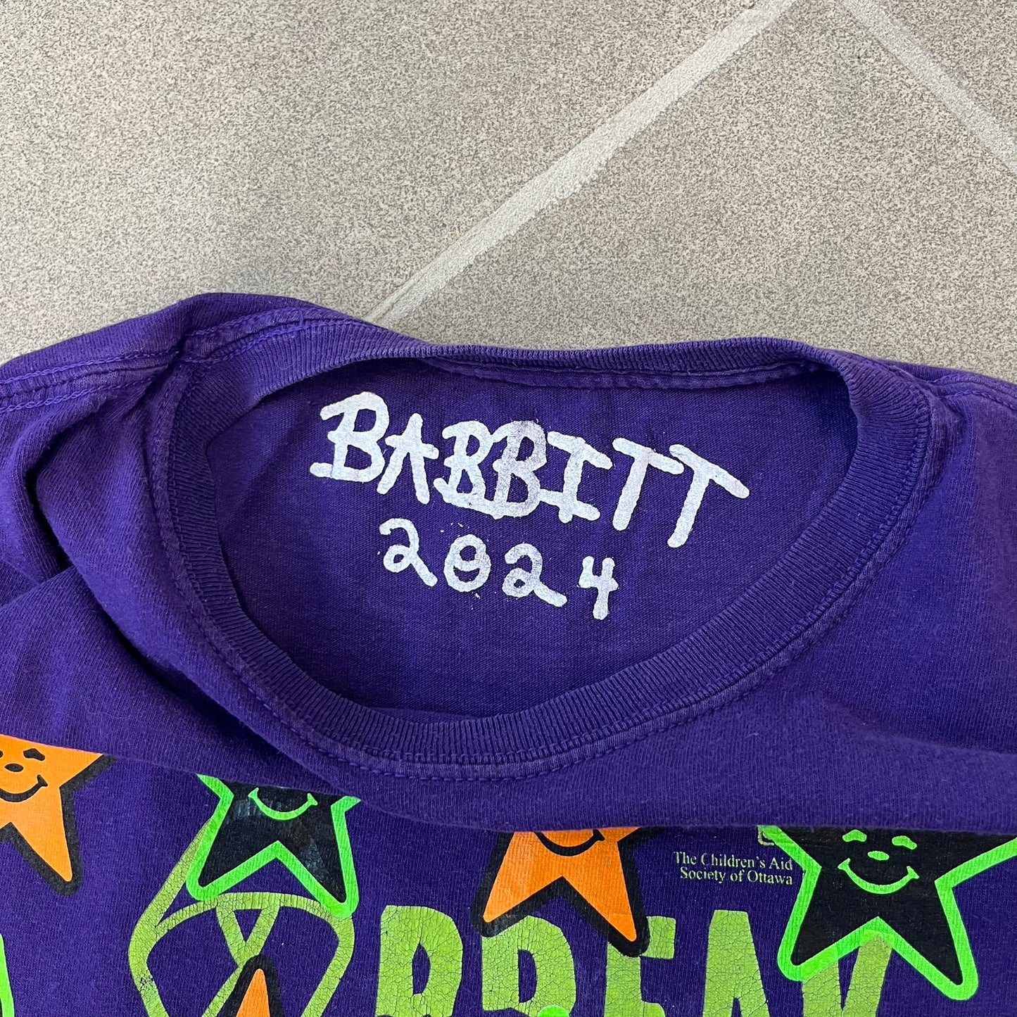 Asspizza 2024 Halloween Stars Purple Tee is a purple T-shirt featuring an all-over star pattern with orange and black stars outlined in green. Each star has a smiling face graphic in the center. The shirt includes a Babbitt 2024 neck tag and is printed on a Children’s Aid blank.
