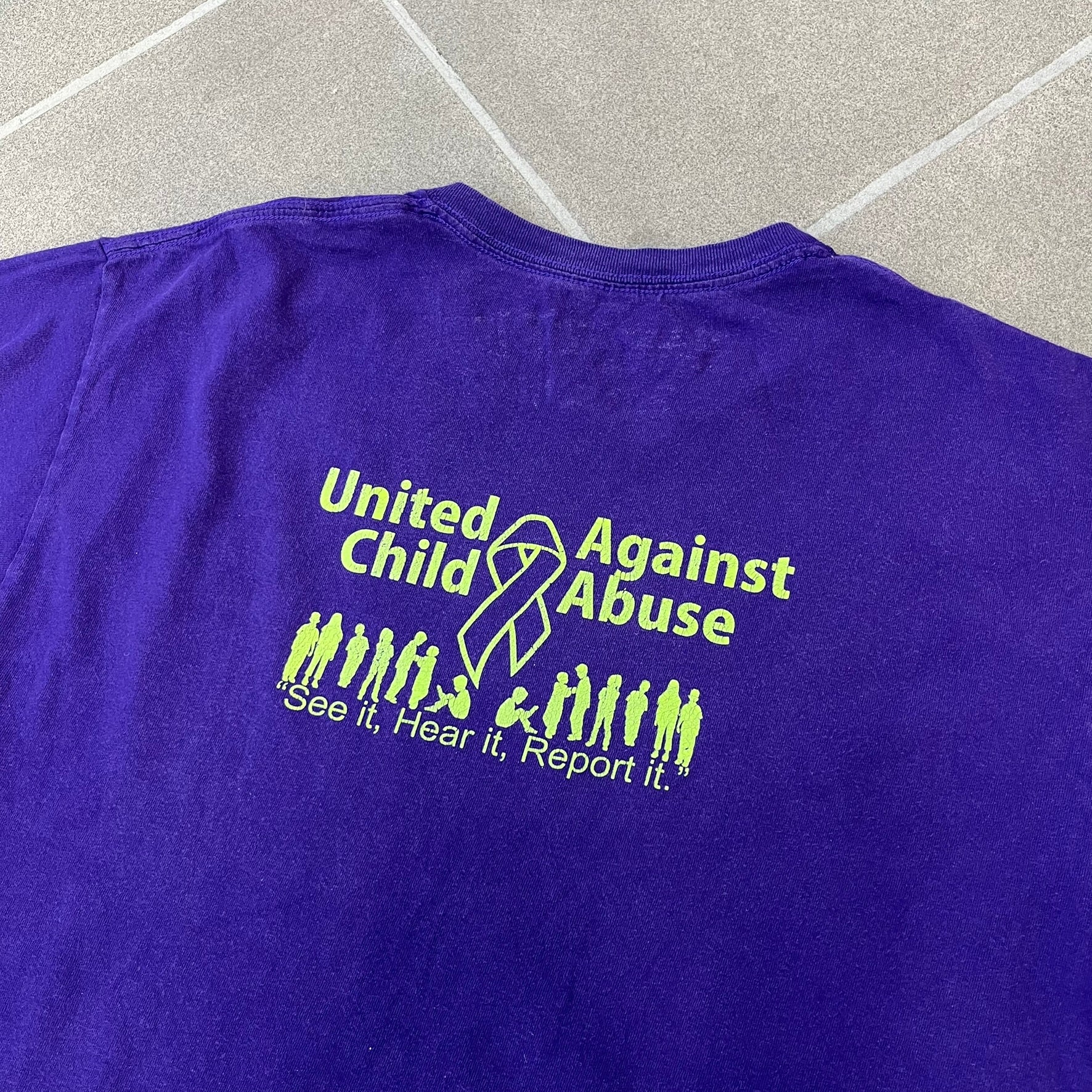 Asspizza 2024 Halloween Stars Purple Tee is a purple T-shirt featuring an all-over star pattern with orange and black stars outlined in green. Each star has a smiling face graphic in the center. The shirt includes a Babbitt 2024 neck tag and is printed on a Children’s Aid blank.
