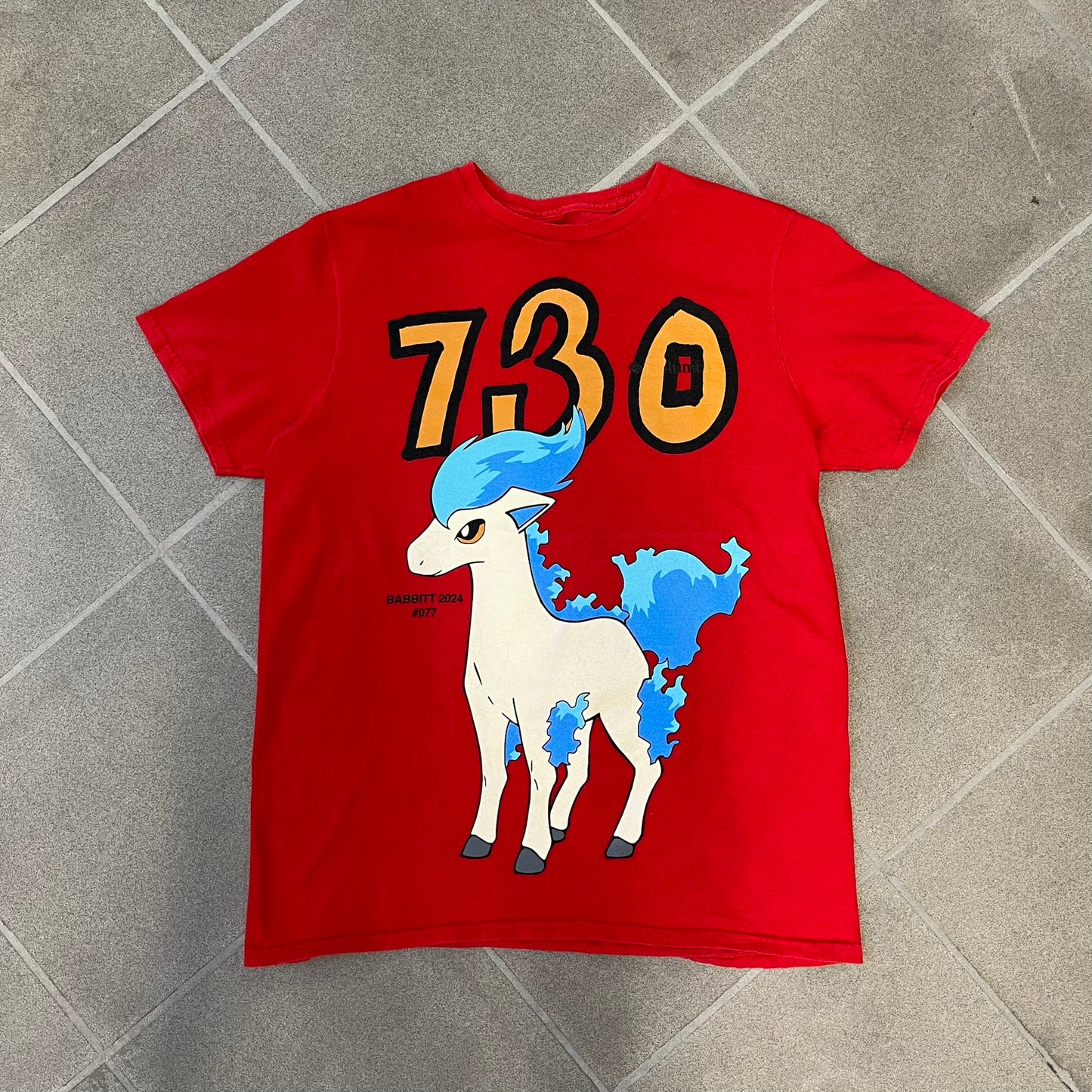 Asspizza 2024 Shiny Ponyta Red Tee is a red T-shirt featuring a shiny Ponyta graphic on the front with a yellow “730” number printed above. Next to the Ponyta graphic is a small black “Babbitt 2024 #077” text detail. The shirt is printed on a Colombia blank.
