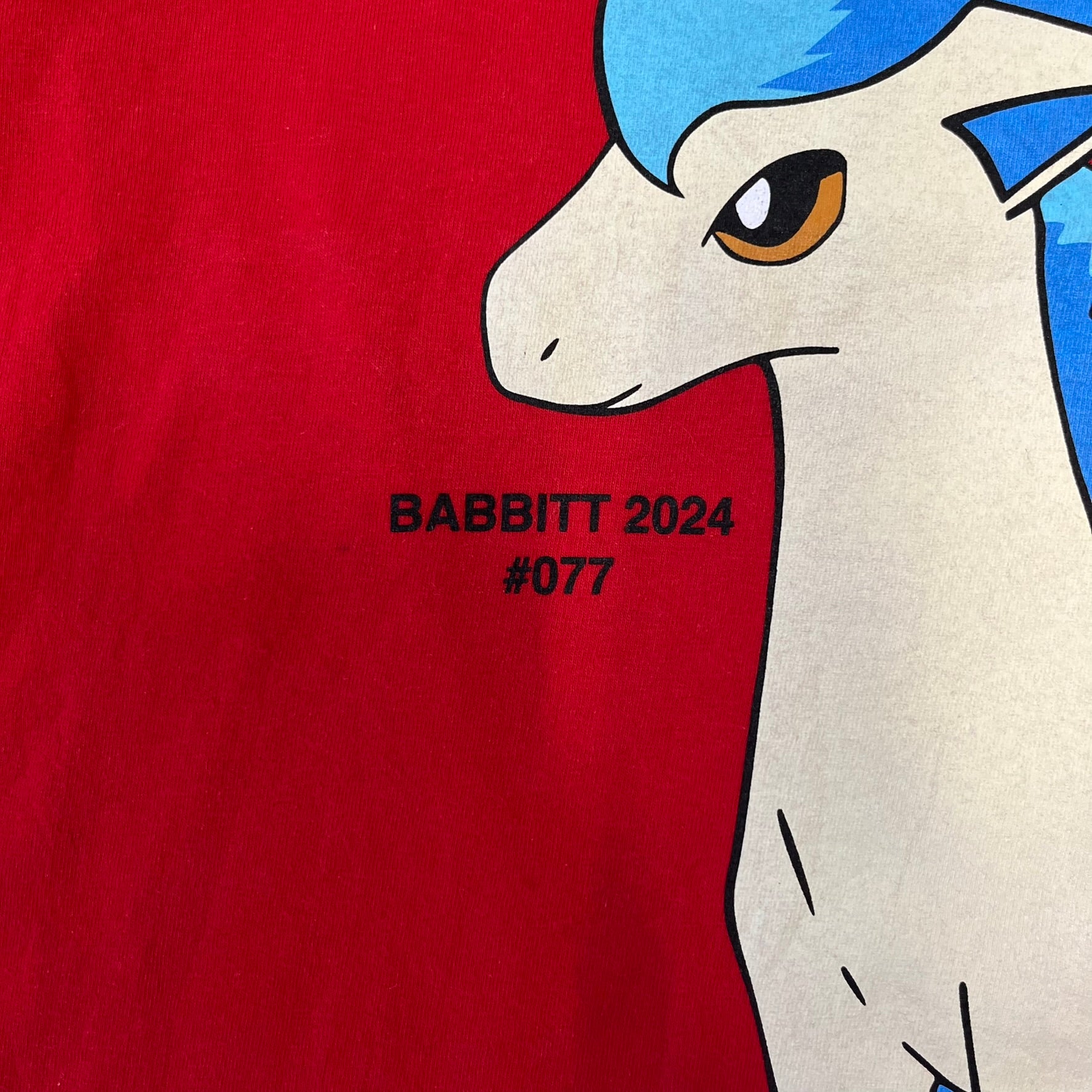 Asspizza 2024 Shiny Ponyta Red Tee is a red T-shirt featuring a shiny Ponyta graphic on the front with a yellow “730” number printed above. Next to the Ponyta graphic is a small black “Babbitt 2024 #077” text detail. The shirt is printed on a Colombia blank.
