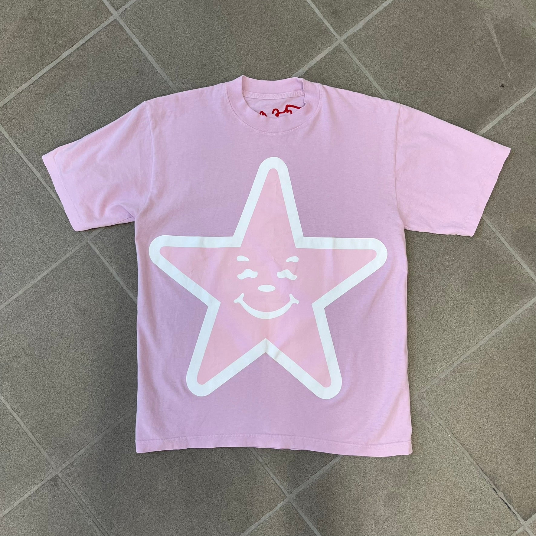 Asspizza 2025 Pink Star Tee is a dark pink T-shirt featuring a large light pink star graphic on the front. The star has a white outline and a white Happy Asspizza face design in the center. The shirt includes a red scribbled 2025 neck tag.
