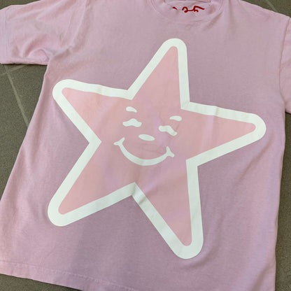 Asspizza 2025 Pink Star Tee is a dark pink T-shirt featuring a large light pink star graphic on the front. The star has a white outline and a white Happy Asspizza face design in the center. The shirt includes a red scribbled 2025 neck tag.
