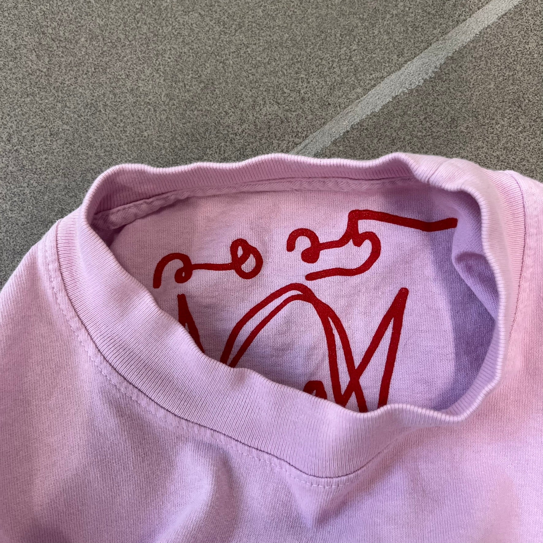 Asspizza 2025 Pink Star Tee is a dark pink T-shirt featuring a large light pink star graphic on the front. The star has a white outline and a white Happy Asspizza face design in the center. The shirt includes a red scribbled 2025 neck tag.
