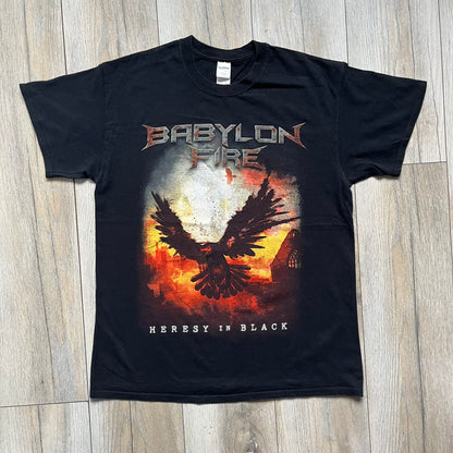 Babylon Fire 2017 “Heresy In Black” Tee is a black T-shirt made on a Gildan blank. The front features the album artwork depicting a fiery landscape with a black bird flying overhead. A stylized metal-inspired “Babylon Fire” logo is printed above the graphic, with “Heresy In Black” text below. Babylon Fire was an alternative rock band active until 2019.