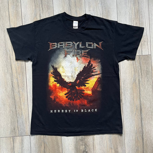 Babylon Fire 2017 “Heresy In Black” Tee is a black T-shirt made on a Gildan blank. The front features the album artwork depicting a fiery landscape with a black bird flying overhead. A stylized metal-inspired “Babylon Fire” logo is printed above the graphic, with “Heresy In Black” text below. Babylon Fire was an alternative rock band active until 2019.