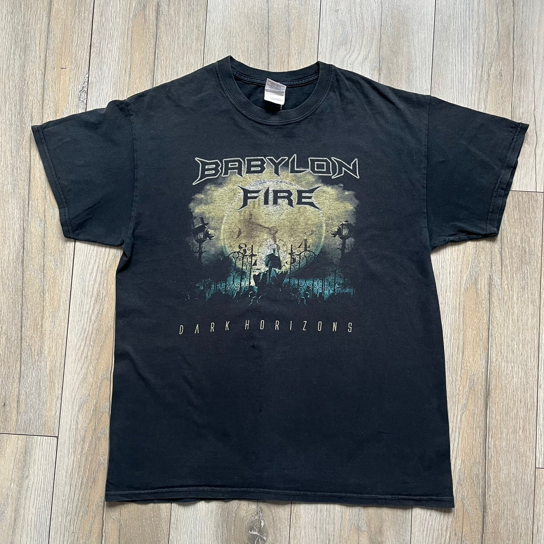 Babylon Fire 2012 “Dark Horizons” Tee is a washed black T-shirt made on a Gildan blank. The front features the album artwork for Dark Horizons, depicting a graveyard scene with a moon and clock. “Babylon Fire” text is printed at the top of the graphic, with “Dark Horizons” text below. Babylon Fire was an alternative rock band active until 2019.