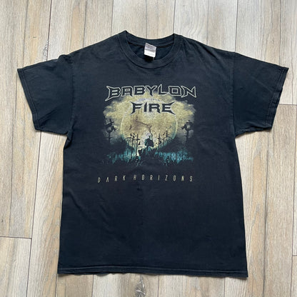Babylon Fire 2012 “Dark Horizons” Tee is a washed black T-shirt made on a Gildan blank. The front features the album artwork for Dark Horizons, depicting a graveyard scene with a moon and clock. “Babylon Fire” text is printed at the top of the graphic, with “Dark Horizons” text below. Babylon Fire was an alternative rock band active until 2019.
