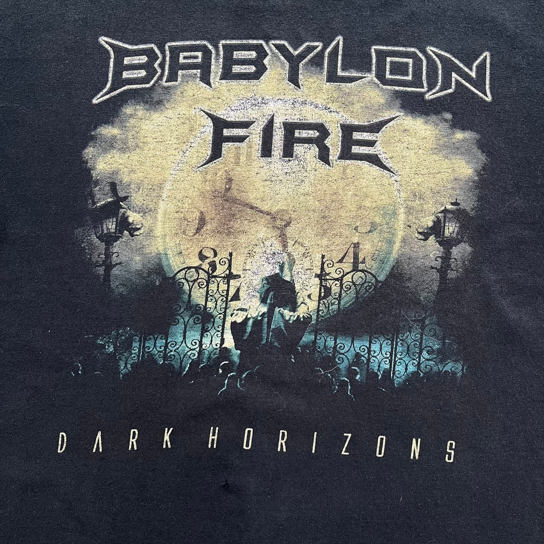 Babylon Fire 2012 “Dark Horizons” Tee is a washed black T-shirt made on a Gildan blank. The front features the album artwork for Dark Horizons, depicting a graveyard scene with a moon and clock. “Babylon Fire” text is printed at the top of the graphic, with “Dark Horizons” text below. Babylon Fire was an alternative rock band active until 2019.