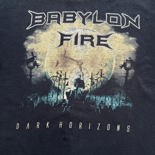 Babylon Fire 2012 “Dark Horizons” Tee is a washed black T-shirt made on a Gildan blank. The front features the album artwork for Dark Horizons, depicting a graveyard scene with a moon and clock. “Babylon Fire” text is printed at the top of the graphic, with “Dark Horizons” text below. Babylon Fire was an alternative rock band active until 2019.