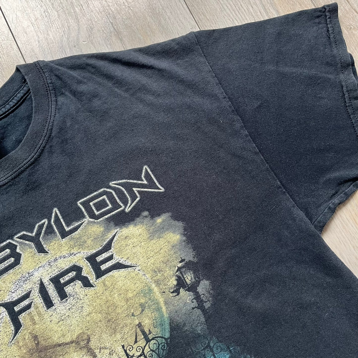 Babylon Fire 2012 “Dark Horizons” Tee is a washed black T-shirt made on a Gildan blank. The front features the album artwork for Dark Horizons, depicting a graveyard scene with a moon and clock. “Babylon Fire” text is printed at the top of the graphic, with “Dark Horizons” text below. Babylon Fire was an alternative rock band active until 2019.