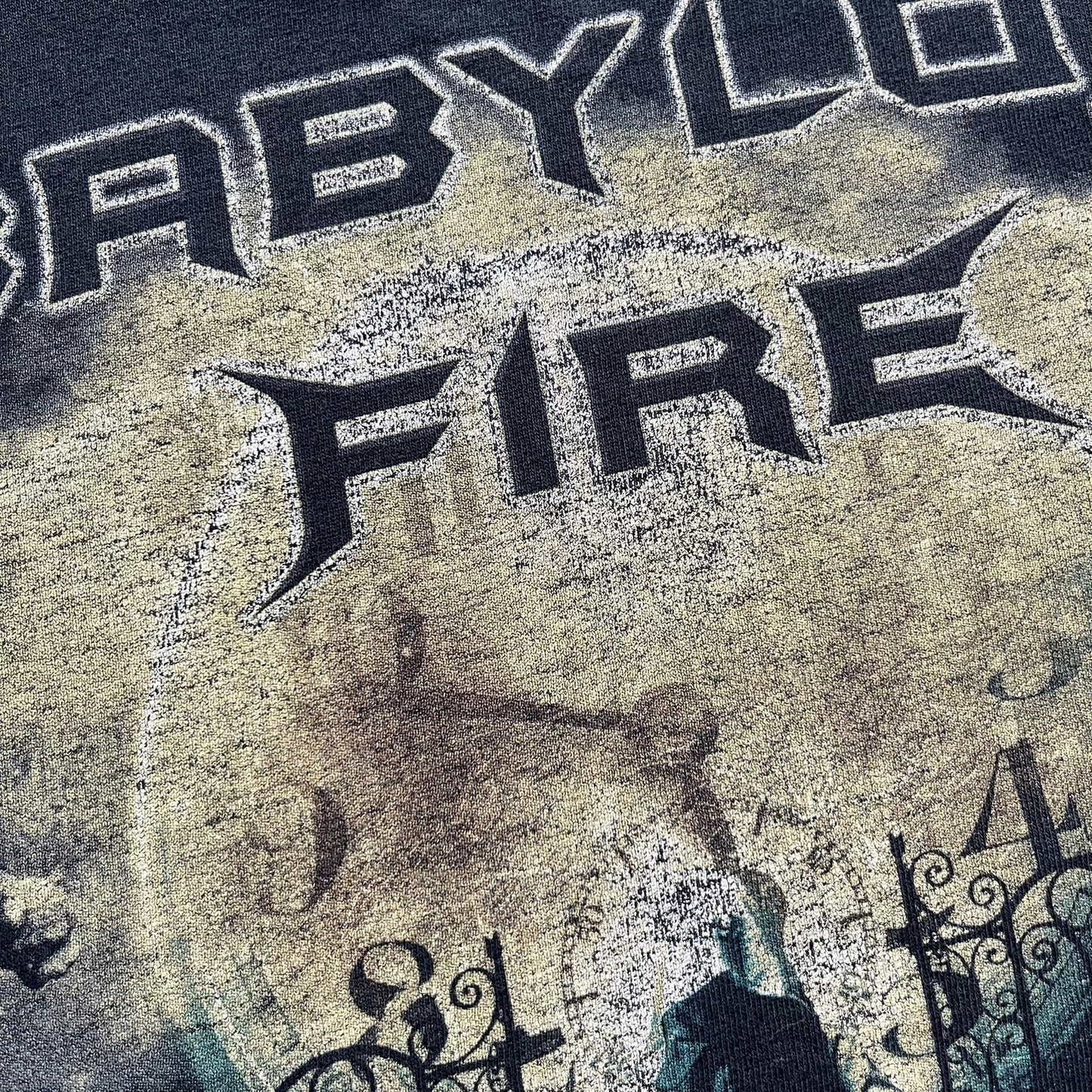 Babylon Fire 2012 “Dark Horizons” Tee is a washed black T-shirt made on a Gildan blank. The front features the album artwork for Dark Horizons, depicting a graveyard scene with a moon and clock. “Babylon Fire” text is printed at the top of the graphic, with “Dark Horizons” text below. Babylon Fire was an alternative rock band active until 2019.