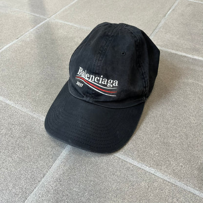 Balenciaga 2017 Political Campaign Baseball Cap features a washed black/smoked grey cotton drill construction with a curved brim. The front has an embroidered white Balenciaga logo above red and white curved stripes, with “2017” embroidered in white underneath. Made from 100% cotton.
