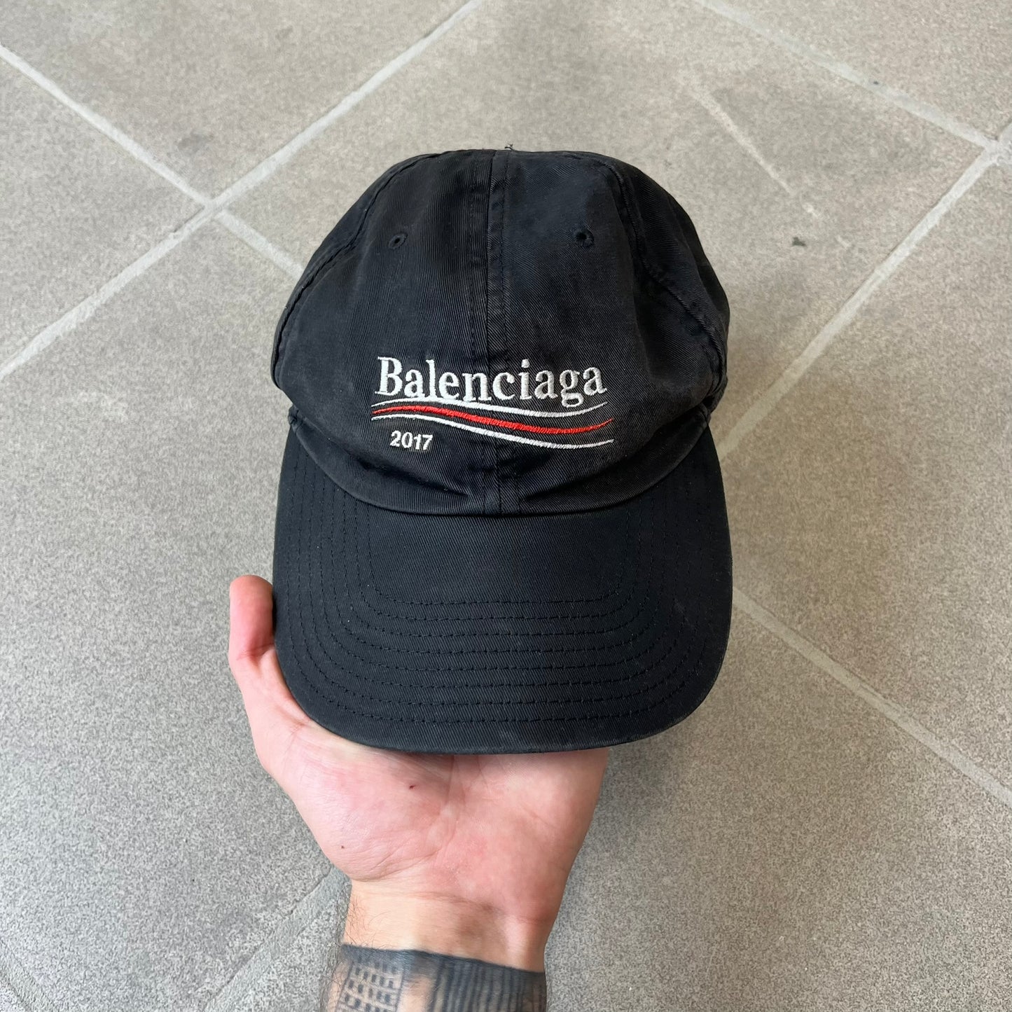 Balenciaga 2017 Political Campaign Baseball Cap features a washed black/smoked grey cotton drill construction with a curved brim. The front has an embroidered white Balenciaga logo above red and white curved stripes, with “2017” embroidered in white underneath. Made from 100% cotton.
