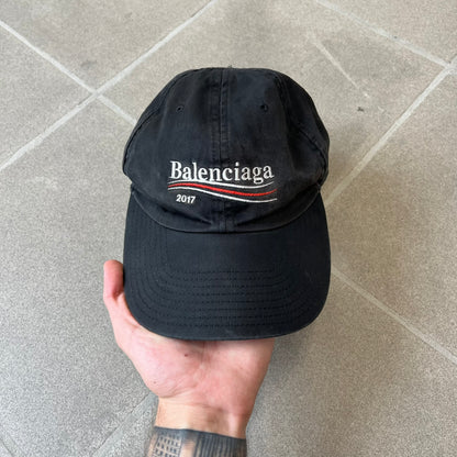 Balenciaga 2017 Political Campaign Baseball Cap features a washed black/smoked grey cotton drill construction with a curved brim. The front has an embroidered white Balenciaga logo above red and white curved stripes, with “2017” embroidered in white underneath. Made from 100% cotton.
