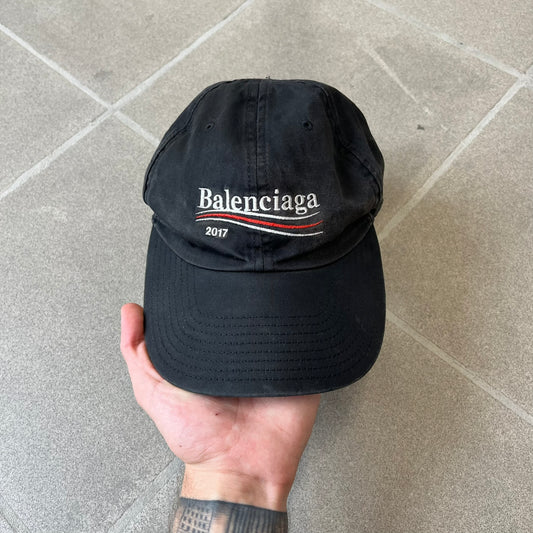 Balenciaga 2017 Political Campaign Baseball Cap features a washed black/smoked grey cotton drill construction with a curved brim. The front has an embroidered white Balenciaga logo above red and white curved stripes, with “2017” embroidered in white underneath. Made from 100% cotton.
