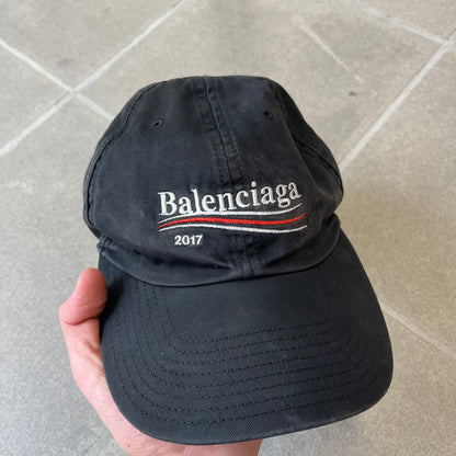 Balenciaga 2017 Political Campaign Baseball Cap features a washed black/smoked grey cotton drill construction with a curved brim. The front has an embroidered white Balenciaga logo above red and white curved stripes, with “2017” embroidered in white underneath. Made from 100% cotton.
