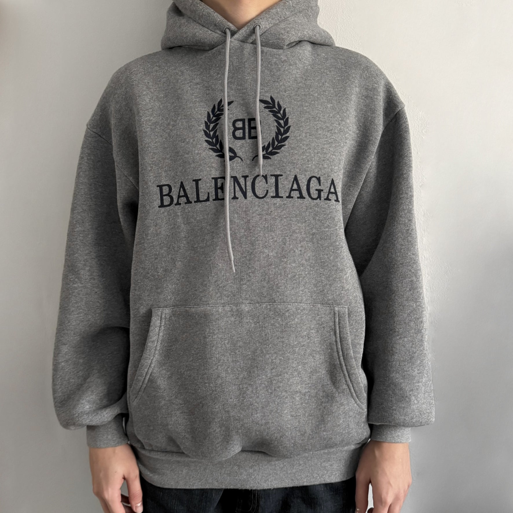 Balenciaga BB Crest Logo Hoodie in Heather Grey, crafted in Portugal from a heavyweight cotton-blend fleece (76% cotton, 24% polyester). The heather grey base features a dark navy BB emblem framed by laurel leaves and the Balenciaga logo text. Finished with dropped shoulders, ribbed edges, and a spacious front pocket, this oversized hoodie exemplifies elevated comfort and craftsmanship.