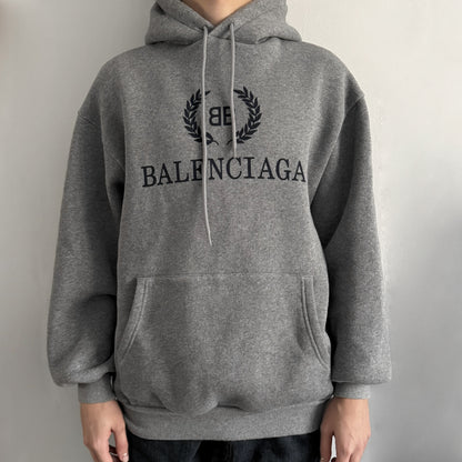 Balenciaga BB Crest Logo Hoodie in Heather Grey, crafted in Portugal from a heavyweight cotton-blend fleece (76% cotton, 24% polyester). The heather grey base features a dark navy BB emblem framed by laurel leaves and the Balenciaga logo text. Finished with dropped shoulders, ribbed edges, and a spacious front pocket, this oversized hoodie exemplifies elevated comfort and craftsmanship.