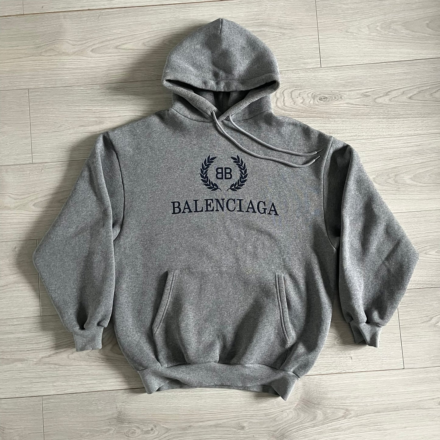 Balenciaga BB Crest Logo Hoodie in Heather Grey, crafted in Portugal from a heavyweight cotton-blend fleece (76% cotton, 24% polyester). The heather grey base features a dark navy BB emblem framed by laurel leaves and the Balenciaga logo text. Finished with dropped shoulders, ribbed edges, and a spacious front pocket, this oversized hoodie exemplifies elevated comfort and craftsmanship.