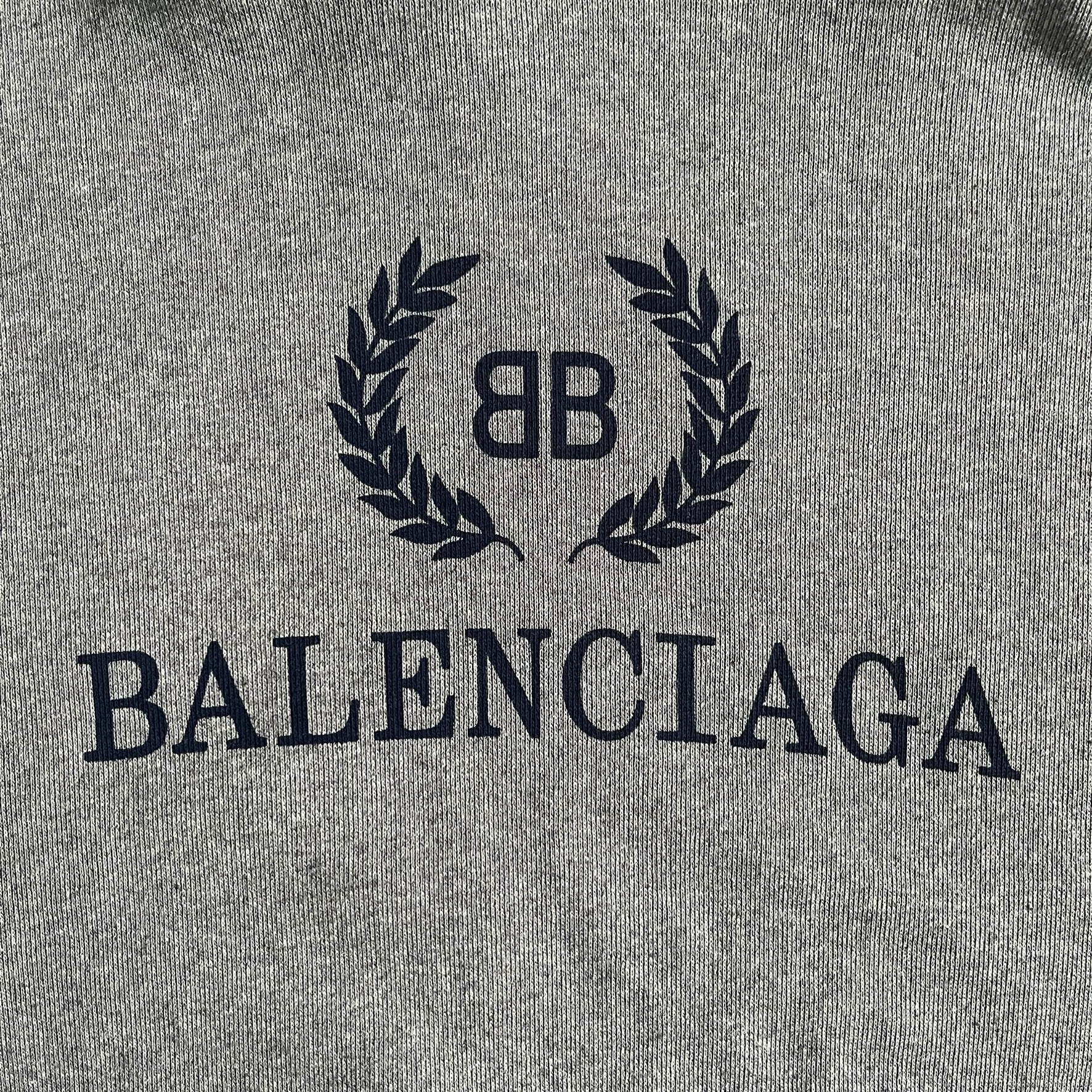 Balenciaga BB Crest Logo Hoodie in Heather Grey, crafted in Portugal from a heavyweight cotton-blend fleece (76% cotton, 24% polyester). The heather grey base features a dark navy BB emblem framed by laurel leaves and the Balenciaga logo text. Finished with dropped shoulders, ribbed edges, and a spacious front pocket, this oversized hoodie exemplifies elevated comfort and craftsmanship.