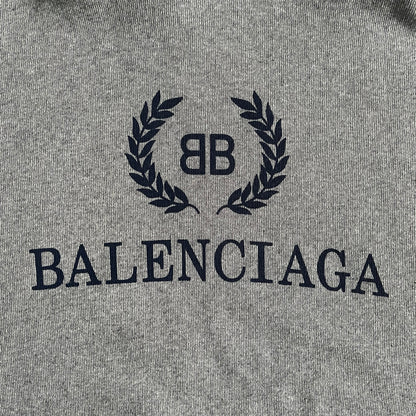 Balenciaga BB Crest Logo Hoodie in Heather Grey, crafted in Portugal from a heavyweight cotton-blend fleece (76% cotton, 24% polyester). The heather grey base features a dark navy BB emblem framed by laurel leaves and the Balenciaga logo text. Finished with dropped shoulders, ribbed edges, and a spacious front pocket, this oversized hoodie exemplifies elevated comfort and craftsmanship.
