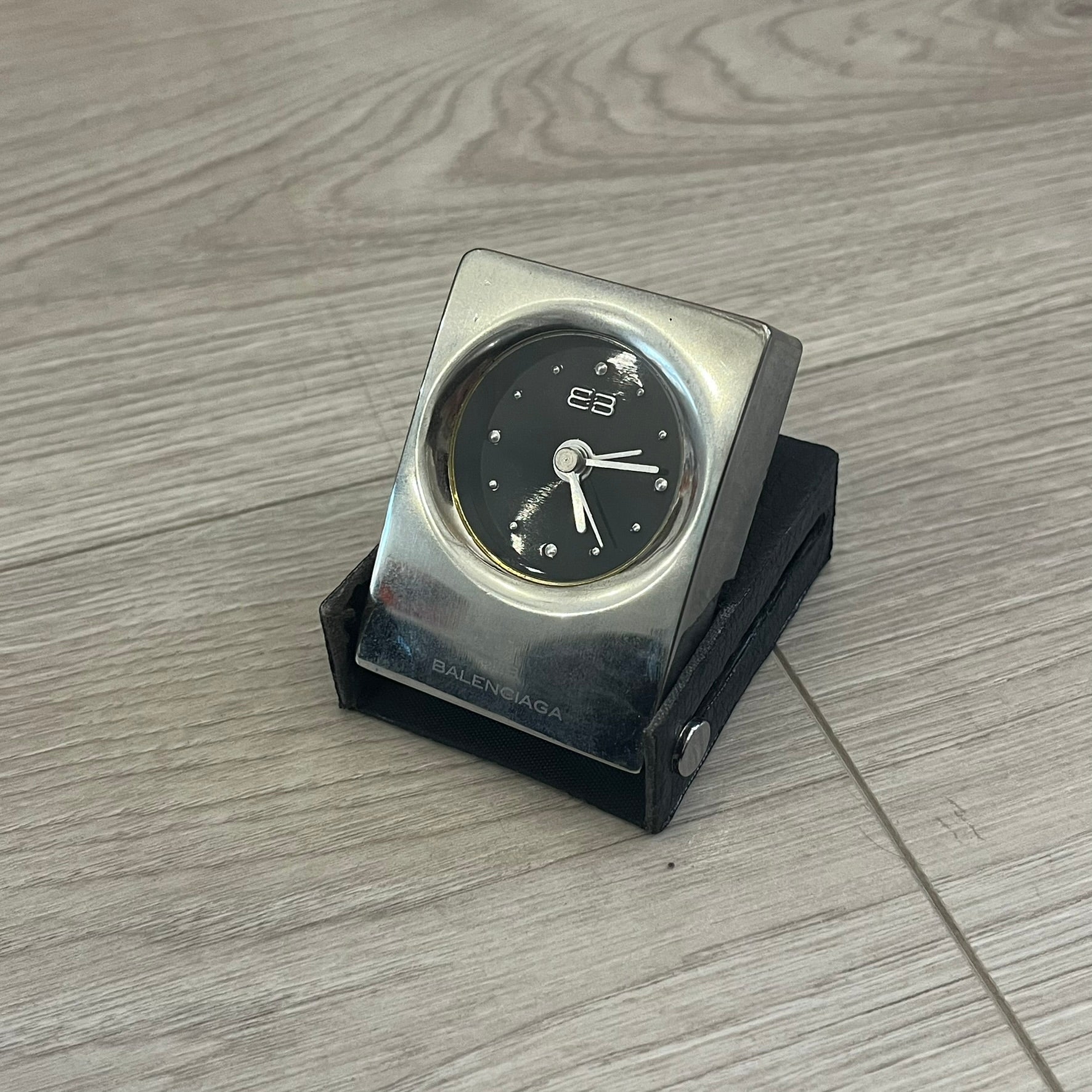 Compact silver-tone metal travel clock with engraved Balenciaga logo. Features a black dial with BB logo and silver-colored hands, plus “Siofor 1000” engraving on the top. Battery-powered and housed in a removable faux leather cover secured by movable side fasteners. Likely produced as a promotional or corporate gift piece.