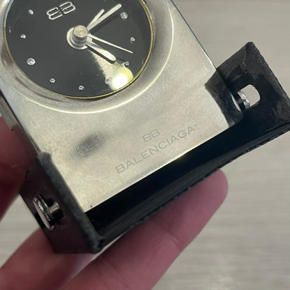 Compact silver-tone metal travel clock with engraved Balenciaga logo. Features a black dial with BB logo and silver-colored hands, plus “Siofor 1000” engraving on the top. Battery-powered and housed in a removable faux leather cover secured by movable side fasteners. Likely produced as a promotional or corporate gift piece.